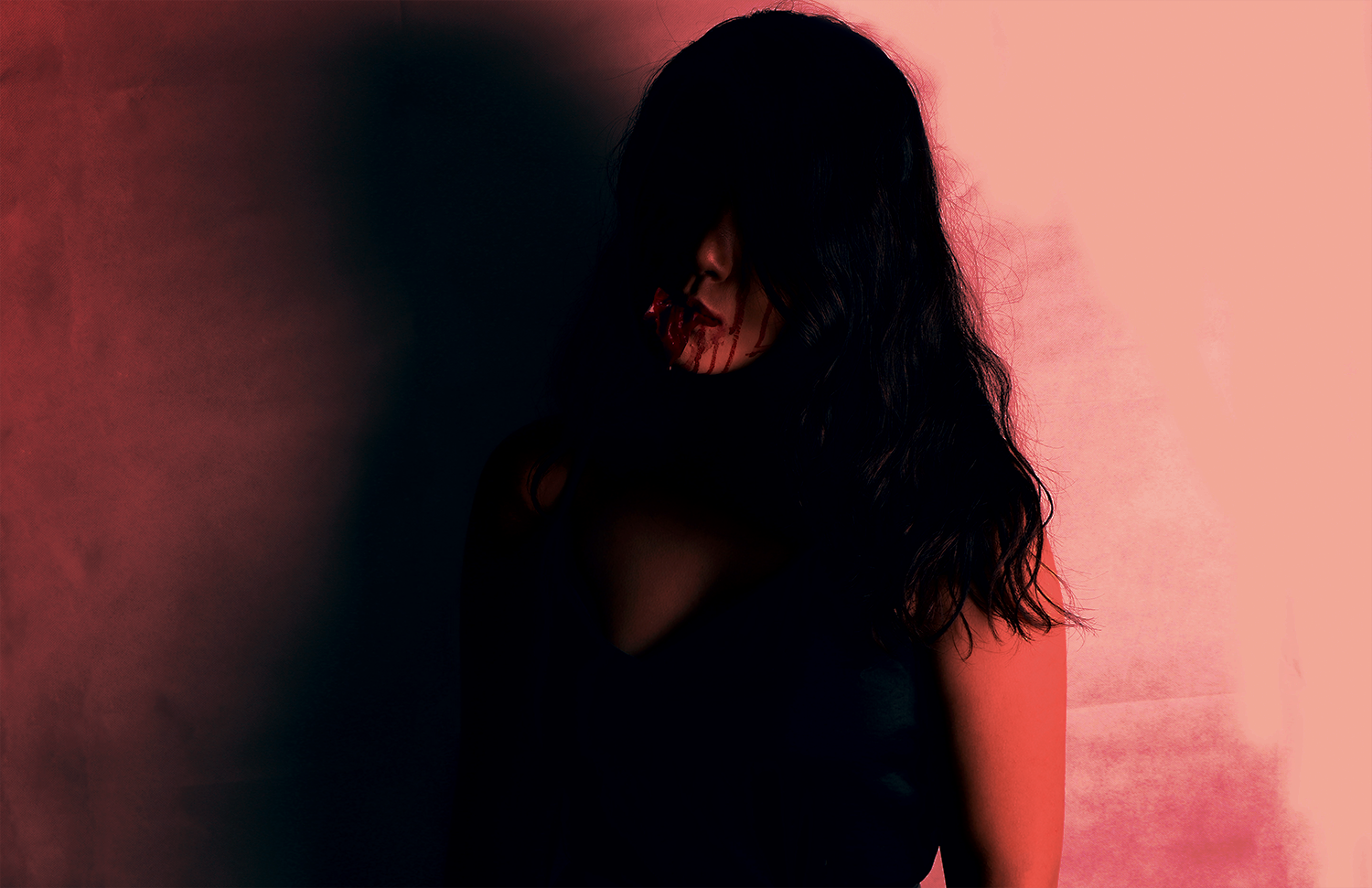 A dark photo of a girl leaning against a wall. Almost all of her face is in shadow with only her mouth visible - which appears to be dripping blood