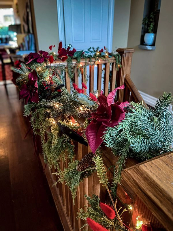 Evergreen boughs and pointsettias wrapped around the top of a stairway railing, interwoven with string lights