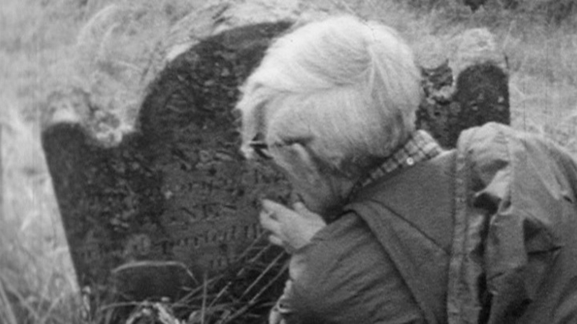 Movie still from The Hungry Ghost showing the protagonist brushing weeds away from the face of a gravestone